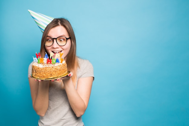 Crazy funny girl in a paper hat and glasses holding a big birthday cake on the blue wall