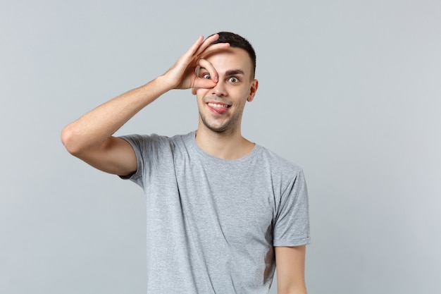 Фото Сумасшедший забавный молодой человек в повседневной одежде показывает язык, держащий руку возле глаз, имитирующих очки или бинокль
