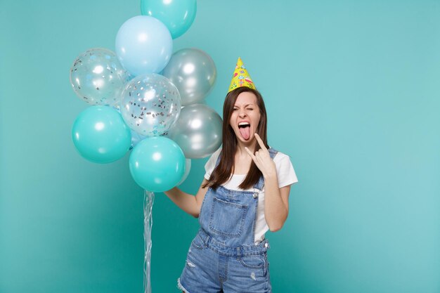 Crazy girl in birthday hat showing tongue horns up heavy metal rock sign celebrating, hold colorful air balloons isolated on blue turquoise background. Birthday holiday party, people emotions concept.