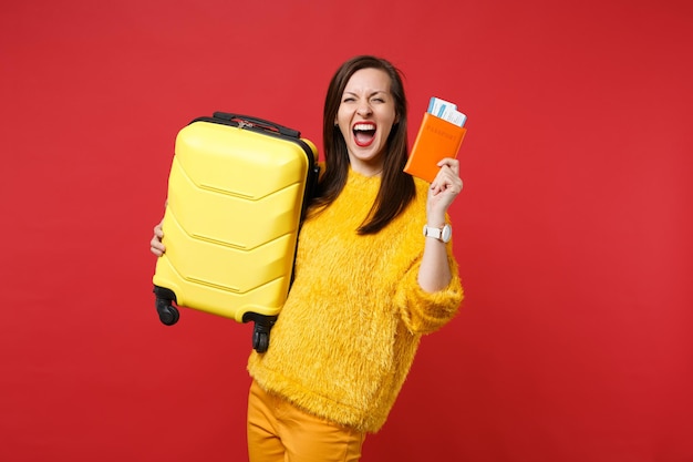 Crazy joyful young woman in yellow fur sweater screaming, holding suitcase, passport boarding pass ticket isolated on red background. People sincere emotions, lifestyle concept. Mock up copy space.