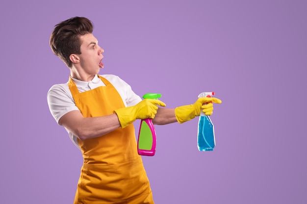 Foto uomo pazzo in grembiule e guanti che grida e spruzza detersivi dalle bottiglie durante la routine di pulizia su sfondo viola