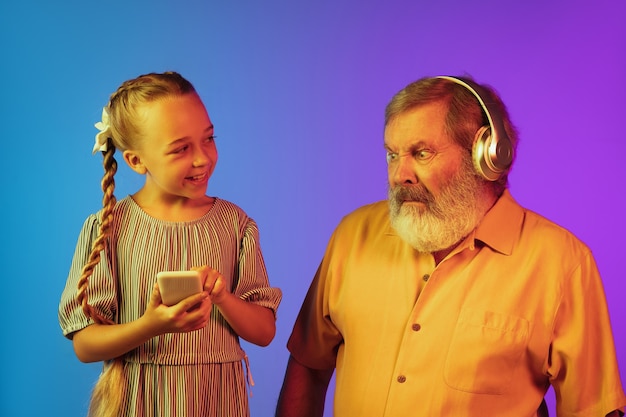 Crazy music of young generation. Senior man spending happy time with granddaughter in neon. Joyful elderly lifestyle, family, childhood, tech concept. Using headphones, smartphone. Copyspace.