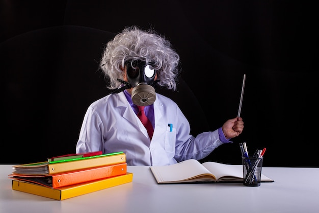 Crazy science teacher in white coat with unkempt hair sitting at the desk  wears a gas mask