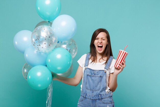 Crazy screaming young woman blinking, holding plastic cup of cola or soda celebrating with colorful air balloons isolated on blue turquoise background. Birthday holiday party, people emotions concept.