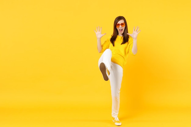 Crazy screaming young woman in casual clothes, heart glasses showing palms, kicking something isolated on yellow orange wall background. People sincere emotions lifestyle concept. Mock up copy space.