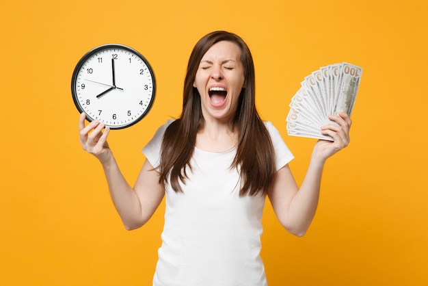 Crazy screaming young woman in white casual clothes holding round clock, fan of cash money in dollar banknotes isolated on yellow orange wall background. People lifestyle concept. Mock up copy space.