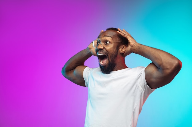 Crazy shocked. African-american young man's portrait on gradient studio background in neon. Beautiful male model in casual style, white shirt. Concept of human emotions, facial expression, sales, ad.