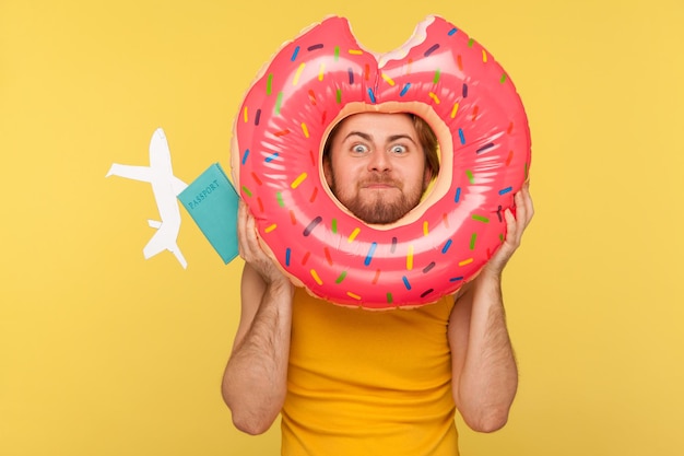Crazy tourist guy in undershirt looking through donut rubber ring with big eyes surprised expression, holding passport document and airplane mockup. indoor studio shot isolated on yellow background