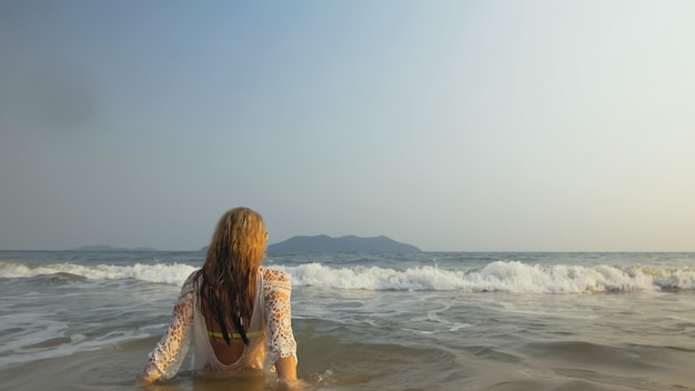 Crazy Woman in a White Tunic on the Beach near the Stormy Sea Cheerful Funny Charming Blonde with Wet Curly Hair in Sunglasses Smiling Happy Girl Tourist Lies in the Water has Fun Back View
