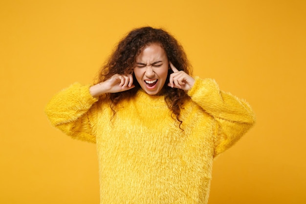 Photo crazy young african american girl in fur sweater posing isolated on yellow orange background. people lifestyle concept. mock up copy space. screaming, keeping eyes closed, covering ears with fingers.