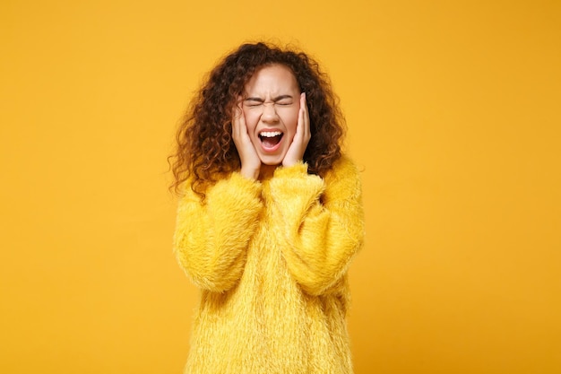 Crazy young african american girl in fur sweater posing isolated on yellow orange wall background. People lifestyle concept. Mock up copy space. Screaming keeping eyes closed, putting hands on cheeks.