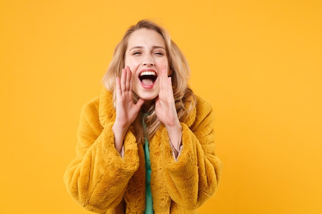 Crazy young blonde woman girl in yellow fur coat posing isolated on orange background studio portrait. People emotions lifestyle concept. Mock up copy space. Screaming with hands gesture near mouth.