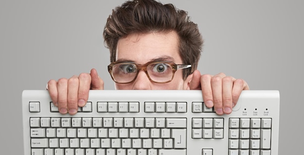 灰色の背景に対してコンピューターのキーボードの後ろからカメラを見てオタク眼鏡のクレイジー若い男性プログラマー