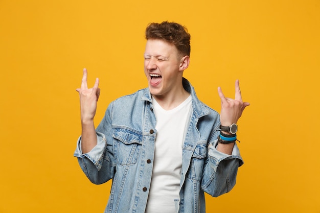 Crazy young man in denim clothes keeping eyes closed, showing horns up gesture, rock-n-roll sign isolated on yellow orange background. People sincere emotions, lifestyle concept. Mock up copy space.