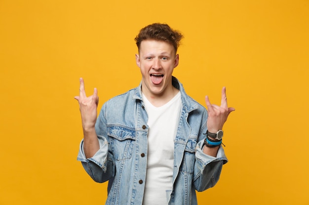 Crazy young man in denim clothes showing tongue, horns up gesture, depicting heavy metal rock sign isolated on yellow orange background. People sincere emotions, lifestyle concept. Mock up copy space.