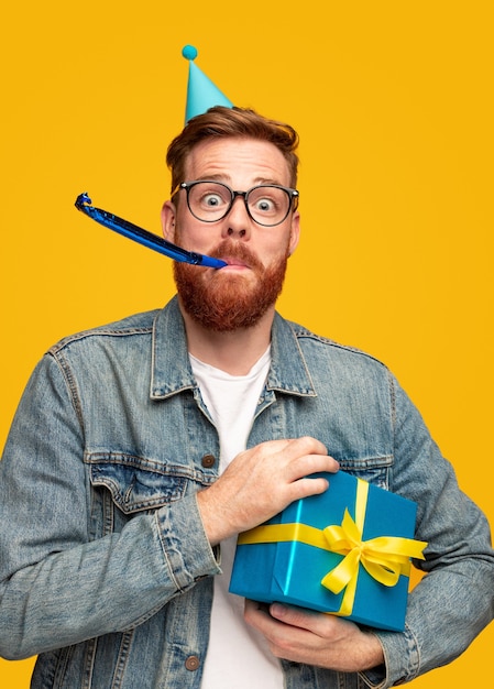 Foto giovane pazzo con la barba allo zenzero che tiene il contenitore di regalo avvolto e che soffia noisemaker durante la festa di compleanno su sfondo giallo