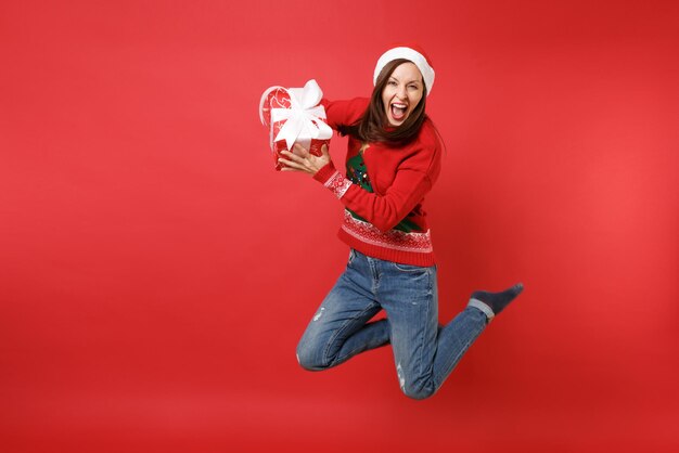 Giovane ragazza pazza di santa in cappello di natale che salta tenendo in mano una scatola rossa con un regalo, urlando presente isolato su sfondo rosso della parete. felice anno nuovo 2019 celebrazione festa concetto. mock up copia spazio.