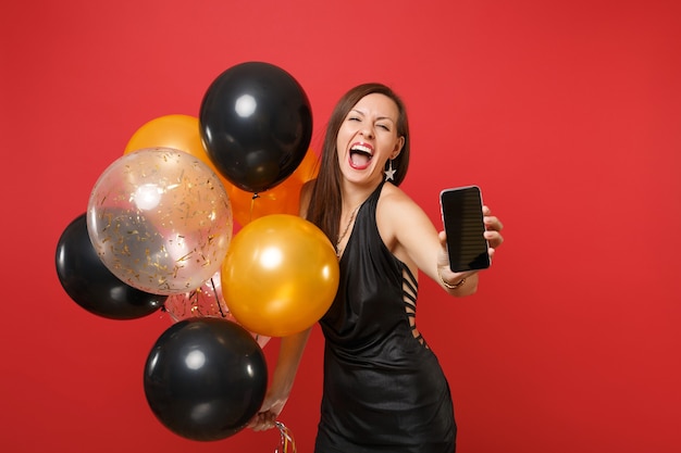 Crazy young woman in black dress screaming celebrating holding mobile phone with blank black empty screen air balloons isolated on red background. Happy New Year birthday mockup holiday party concept.