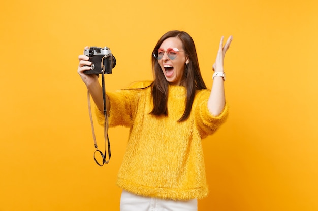 Photo crazy young woman in heart glasses screaming spreading hands doing taking selfie shot on retro vintage photo camera isolated on yellow background. people sincere emotions, lifestyle. advertising area.
