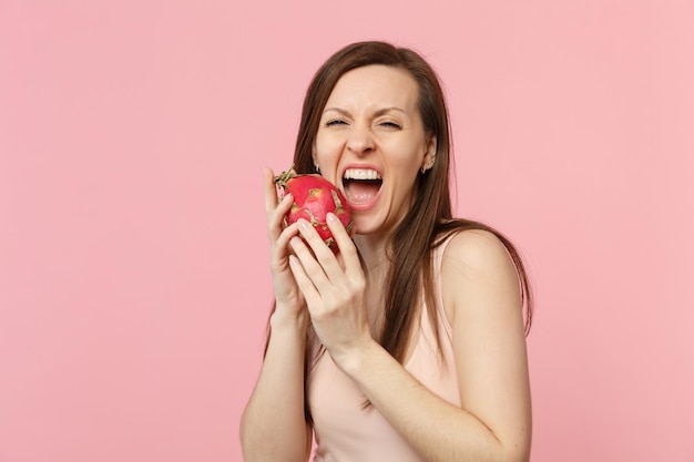 La giovane donna pazza in abiti leggeri tiene, morde la pitahaya fresca matura, il frutto del drago isolato su uno sfondo rosa pastello della parete in studio. persone stile di vita vivido, relax concetto di vacanza. mock up spazio di copia.