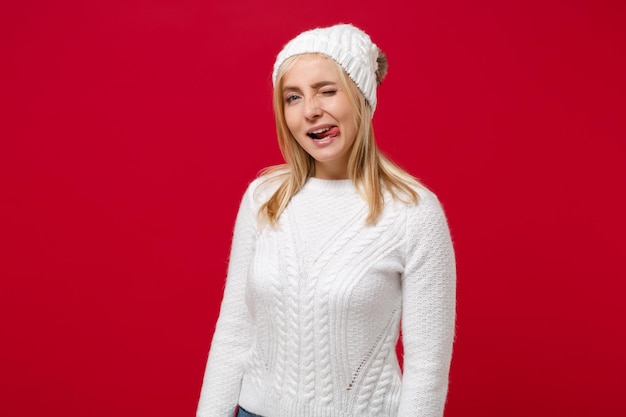 Giovane donna pazza in maglione bianco, cappello isolato su sfondo muro rosso, ritratto in studio. stile di vita sano della moda, emozioni sincere della gente, concetto di stagione fredda. simula lo spazio della copia. mostrare la lingua.