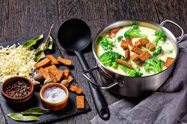 Cream cheese soup with broccoli and rye croutons in a pot on a dark wooden table with spoon and ingredients, horizontal view from above