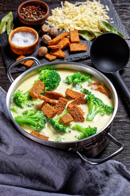 Cream cheese soup with broccoli and rye croutons in a pot on a dark wooden table with spoon and ingredients, vertical view from above