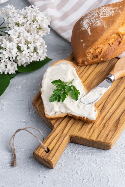 근처에 치즈 나이프와 흰색 라일락이 있는 신선한 바삭한 호밀 빵 조각에 허브를 곁들인 크림 치즈