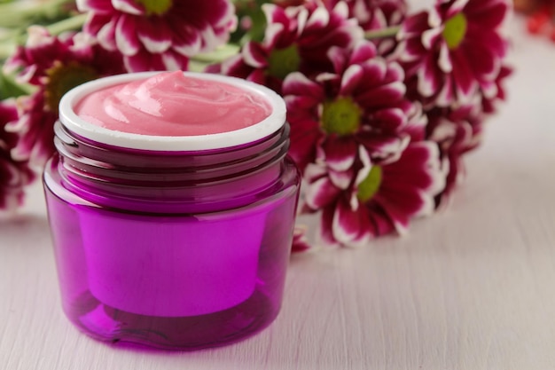 Cream cosmetics for face and body Pink cream and flower in a purple jar on a white wooden table