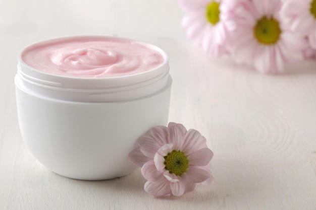 Cream cosmetics for face and body Pink cream and flower in a white jar on a white wooden table