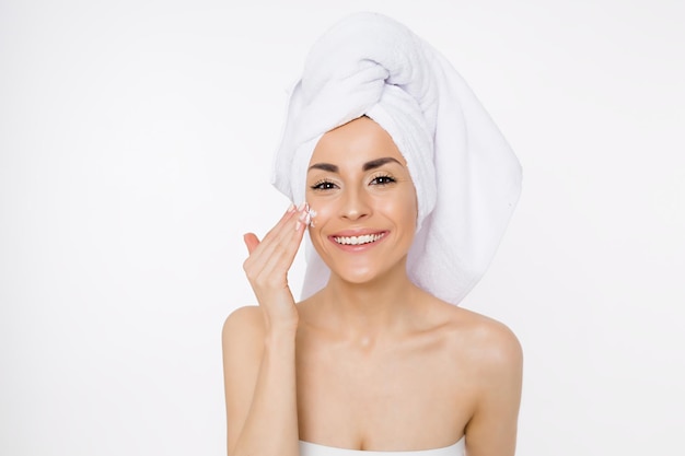 Cream for face A beautiful smiling young woman is applying a thin layer of cream on her face Procedures after shower Girl with a towel on her head