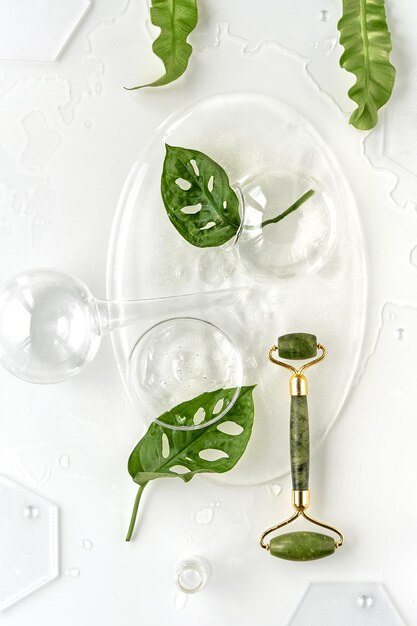 Cream green jade face roller on ice with ice cubes and exotic leaves Monstera Adansonii leaves Minimal flat lay on off white background Facial massage with home made self made moisturizer