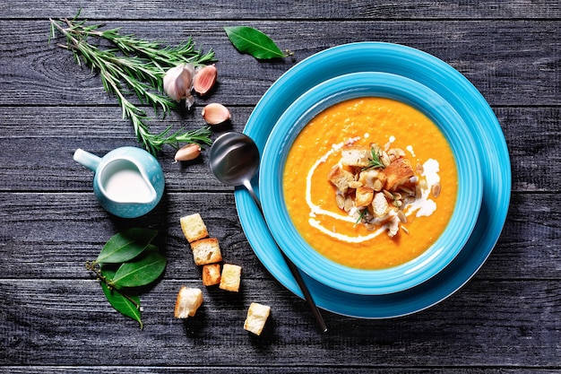 Zuppa di zucca cremosa e confortante piatto autunnale in un piatto blu con crostini di pane e semi di zucca su una superficie di legno scuro con panna, aglio, alloro, rosmarino, vista dall'alto, primo piano