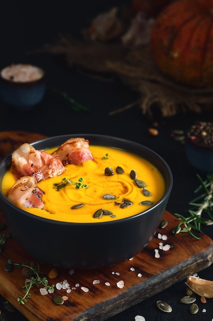 Zuppa di zucca cremosa con fette di pancetta fritta con panna e semi di zucca in ciotola nera sul tavolo di legno nero. primo piano, fuoco selettivo. ingredienti per preparare la zuppa di zucca di stagione in tavola