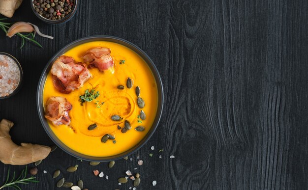 Creamy pumpkin soup with sauteed bacon slices with cream and pumpkin seeds in a black bowl on a black wooden table. Top view with copy space. Ingredients for making pumpkin soup on the table