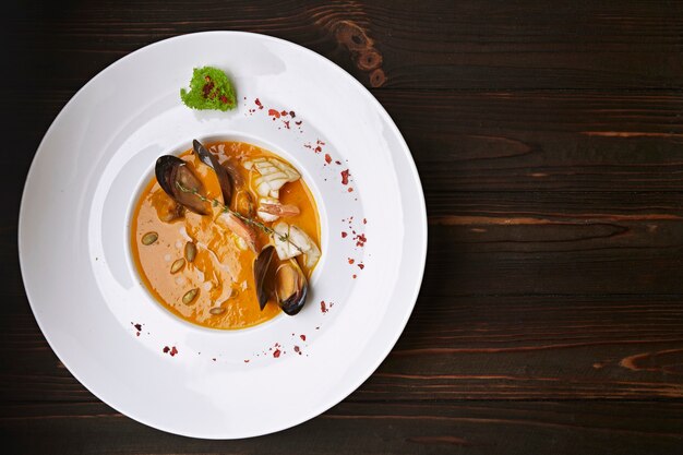 Creamy pumpkin soup with seafood, mussels, shrimps, squids, in a white plate, on a wooden surface