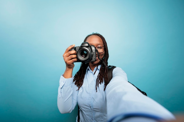 Creatieve amateurfotograaf die foto's maakt met moderne camera voor sociale media. Jonge volwassen Afro-Amerikaanse vrouw die DSLR-apparaat naar de camera richt terwijl ze een foto maakt op een blauwe achtergrond.