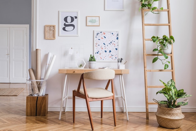 Creatieve compositie van stijlvol scandinavisch kantoor aan huis interieur met posterframes, houten bureau, stoel, planten en accessoires. Neutrale wanden, parketvloer.