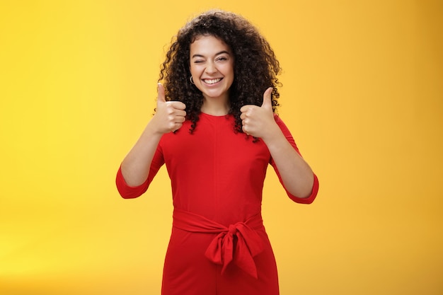 Creatieve en charismatische gelukkige vrolijke vrouw 25s met krullend haar in rode jurk knipogen in goedkeuring en duimen opdagen met een brede glimlach, tevreden positief antwoord geven over gele muur.