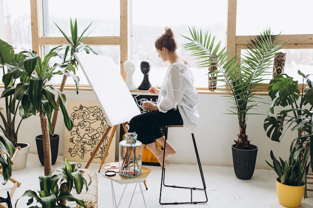 Creatieve jonge vrouw met blote voeten zittend op een stoel en het gebruik van penseel met kleurenpalet voor het schilderen op canvas