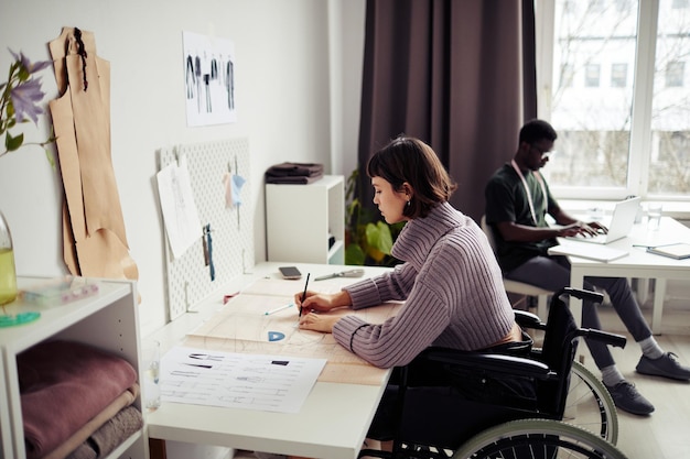 Creatieve jonge vrouw zit in een rolstoel aan haar bureau in de studio en tekent een naaipatroon