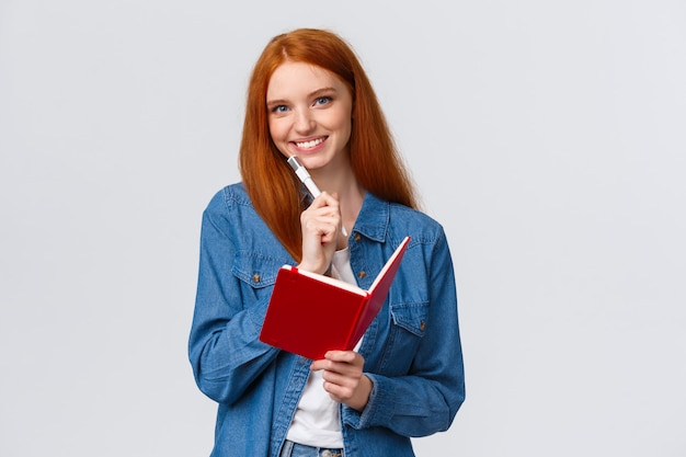 Creatieve schattige goed uitziende roodharige vrouwelijke student, tiener in casual outfit, pen vasthouden en camera glimlachen, notities maken in notitieblok, dagboek schrijven, schema opstellen dat wit staat