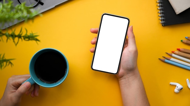 Creatieve stijlvolle gele werkruimtetafel met vrouwelijke mok tijdens het gebruik van het bovenaanzicht van de smartphone