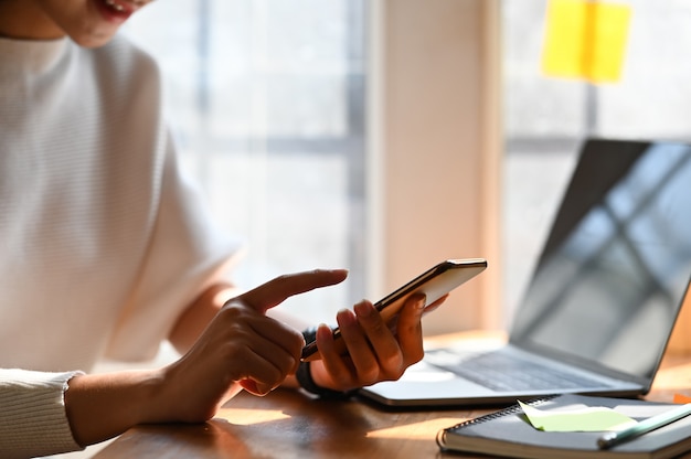 Creatieve vrouw in witte katoenen kledingszitting bij de moderne houten lijst en houdend smartphone in hand vooraan computerlaptop met comfortabele woonkamer as