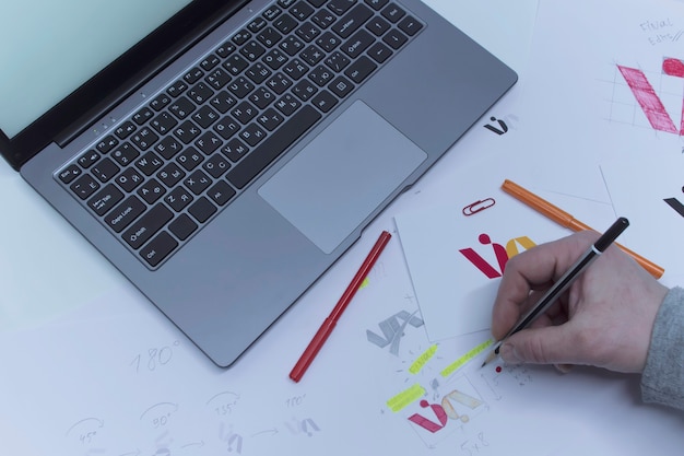 Creatieve werkplek van een grafisch ontwerper. een man op kantoor ontwikkelt een logo op tafel tegen de achtergrond van gedrukte schetsen en een laptop.