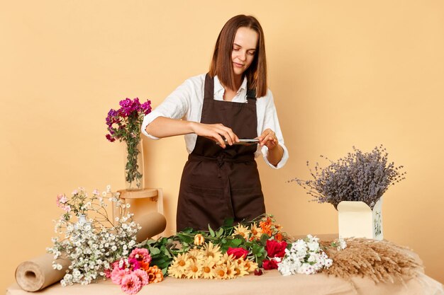 Foto creazione di contenuti per il blog di lavoro bella donna fiorista che fotografa un arrangiamento floreale lavorando con gli ordini in uno studio di design floreale isolato su uno sfondo beige