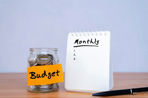 Creating a monthly budget Glass jar with coins and an inscription budget Use a pen to write monthly expenses on a notebook