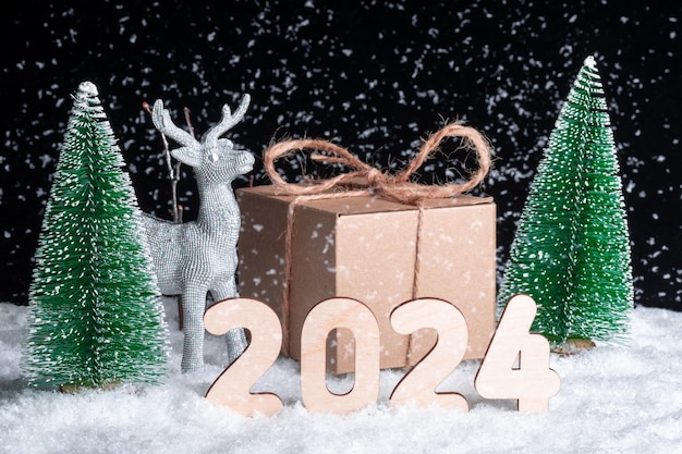 Foto cartolina di natale 2024 creativa un giocattolo splendente di cervo d'argento e una confezione regalo nella neve nella foresta tra gli alberi di natale e numeri di legno 2024 di notte l'aiutante delle renne di babbo natale felice anno nuovo