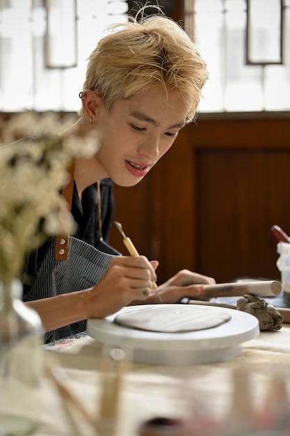 A creative Asian gay man pottery artist holding a sculpting tool making patterns on raw clay