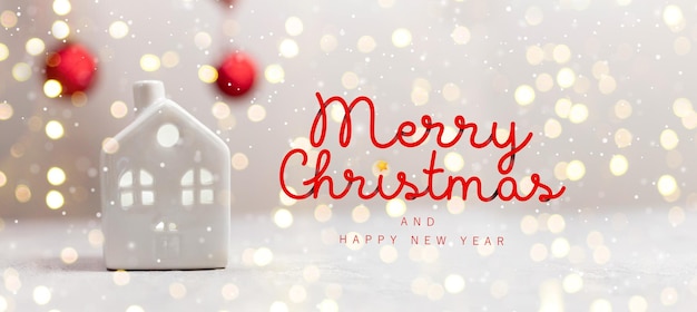 Creative Christmas small toy model house and red balls on white table with light blurred wall background. Holiday template horizontal composition with Merry Christmas text